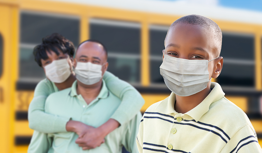 A child wearing a mask in front of a school bus, with his parents in the background